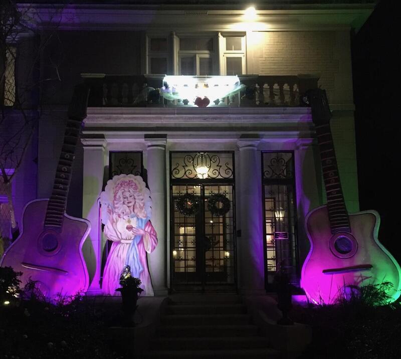 This is a picture of a heavily decorated house . A yellow sign stuck in the lawn out front reads "Vaccine, Vaccine, Vaccine, Vacciiiine!" A giant acoustic guitar sits by the entrance to the home, next to a cardboard cutout of a woman in a flowing pink and purple robe. 