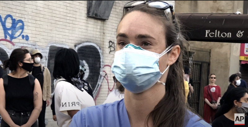 This is a picture of a woman wearing several face masks and a pair of sunglasses looking into the camera. Several other people wear face masks in the background, which appears to be on a city street. 