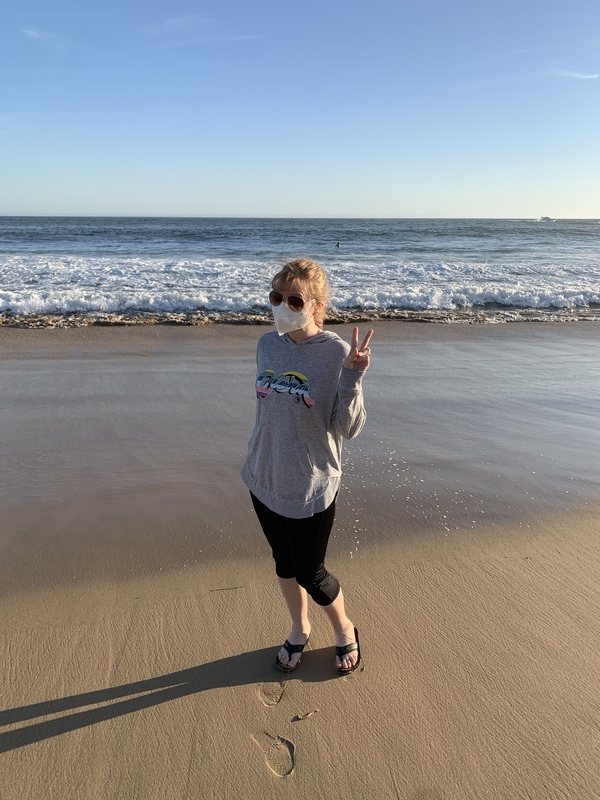 This is a picture of a woman wearing a face mask and sunglasses at the beach, posing for a photo by making a peace sign with one hand. 