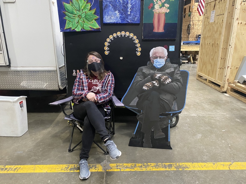This is a picture of a woman who is sitting in a chair wearing a face mask, striking a pose that imitates the pose being taken by the Bernie Sanders cardboard cutout set in the chair next to her. 