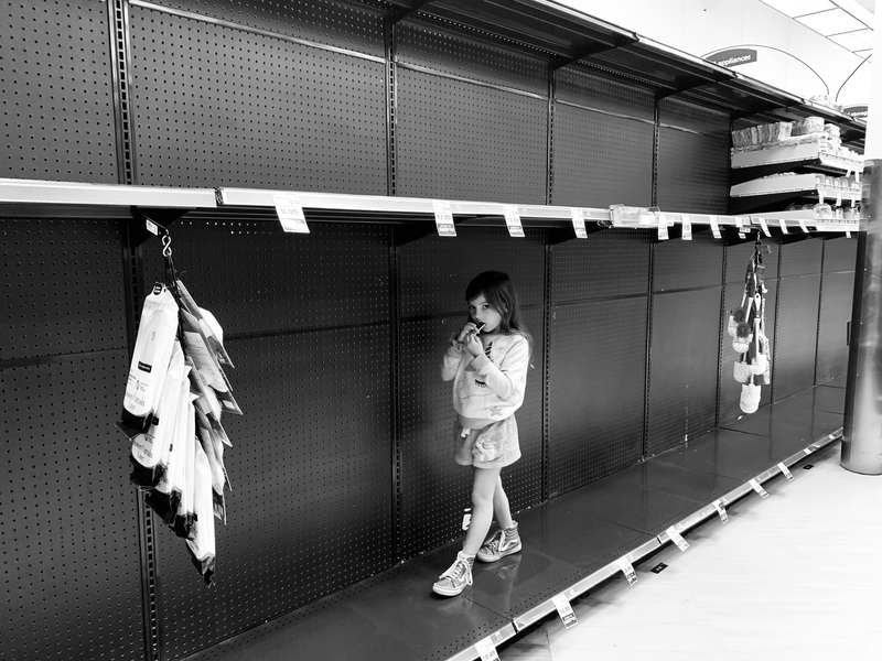 This is a black and white picture of a young girl who is walking on some empty shelves at a store. 