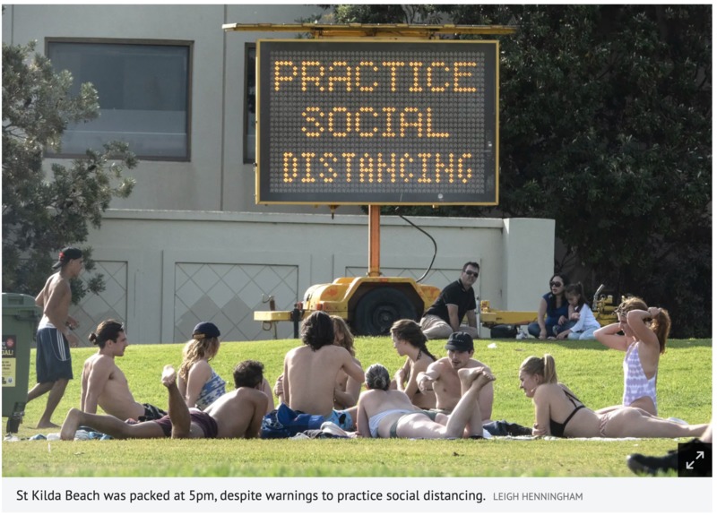 A field of grass has multiple groups of people having picnics and sun bathing. There's an electric sign behind the multiple groups of people that says: PRACTICE SOCIAL DISTANCING. 
