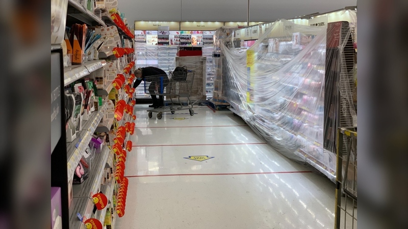 This is a picture of a man in a face mask browsing the shelves of a store, while the refrigerated sections behind him are blocked from customers with plastic wrap. 