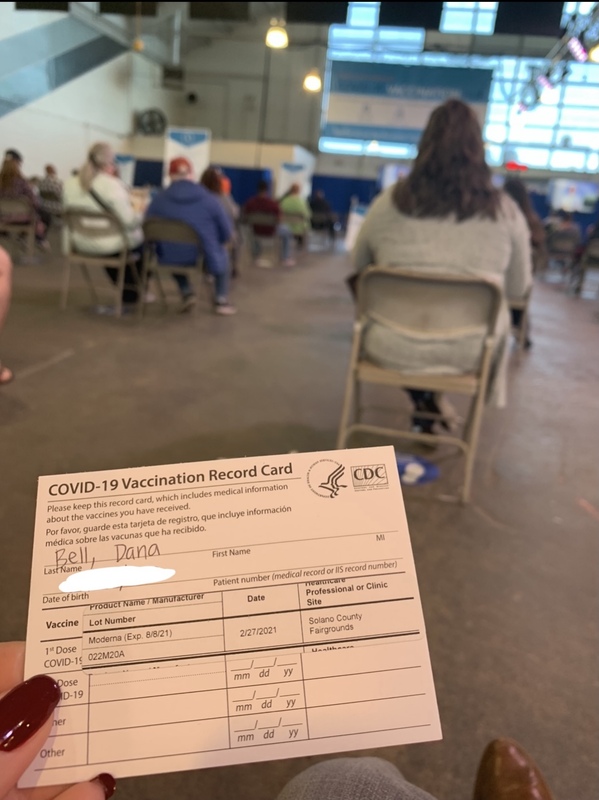 This is a picture taken of a person's vaccine card, with other people waiting to leave the vaccination area in the background. 