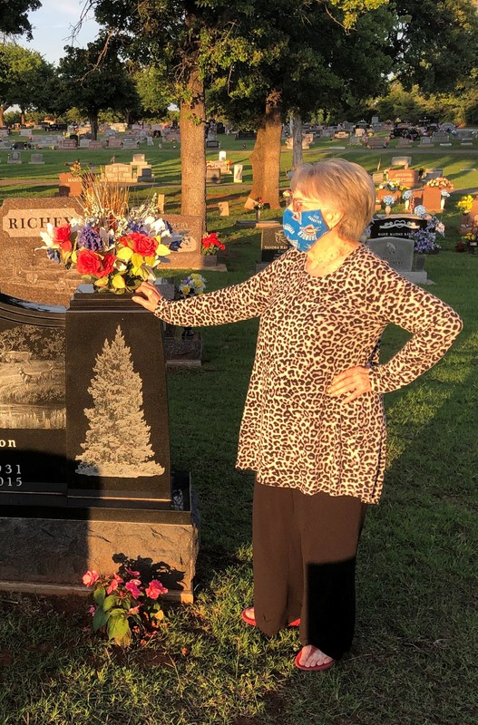 A photo of an older woman wearing a mask and standing by a grave.