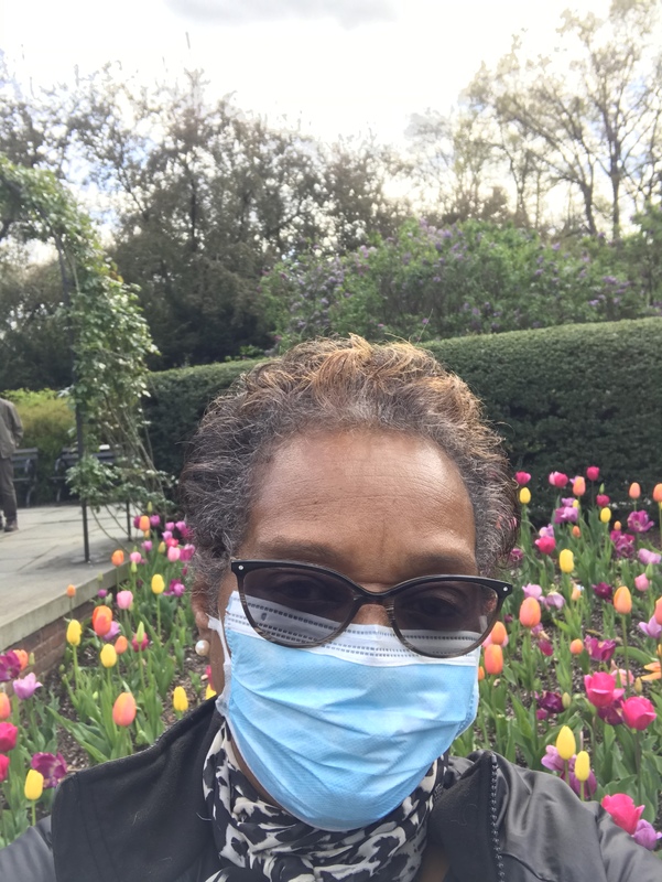 a selfie of a woman outside in a garden in sunglasses and a mask