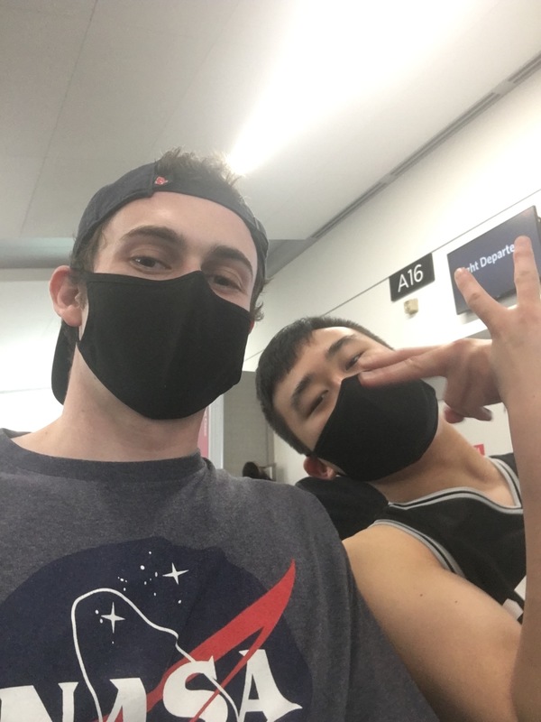 Two men that are posing for a selfie in the airport. The man on the right has a hat on backwards and has his hair sticking out of the hat, his hair is brown. He's wearing a navy shirt with a NASA logo on it. The man on the right is posing and is giving the camera two peace signs and is wearing a black and white tank top. both of the men are wearing black face masks. 