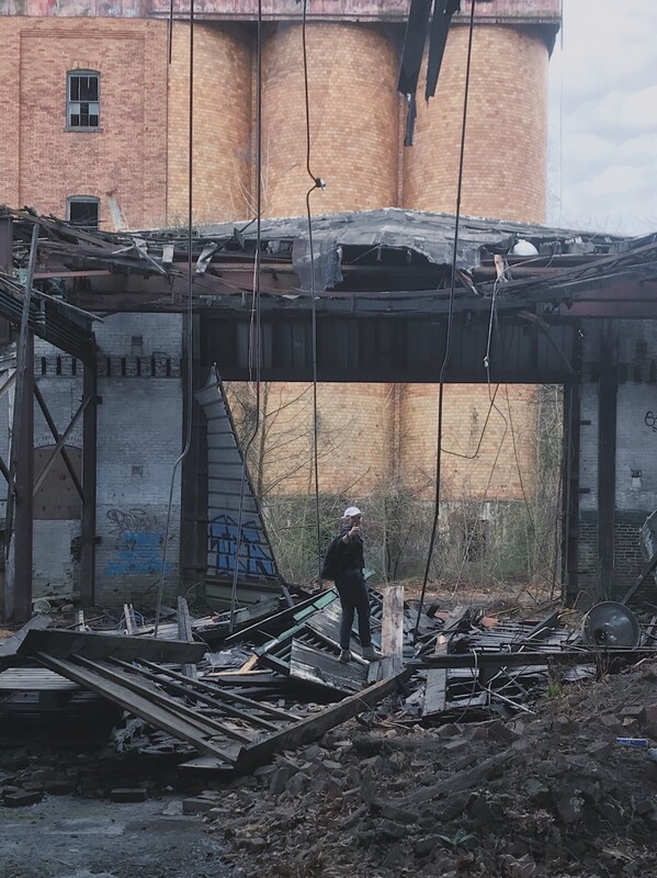 Girl in black clothes and white hat gives "thumbs up" sign while standing in a demolished building.