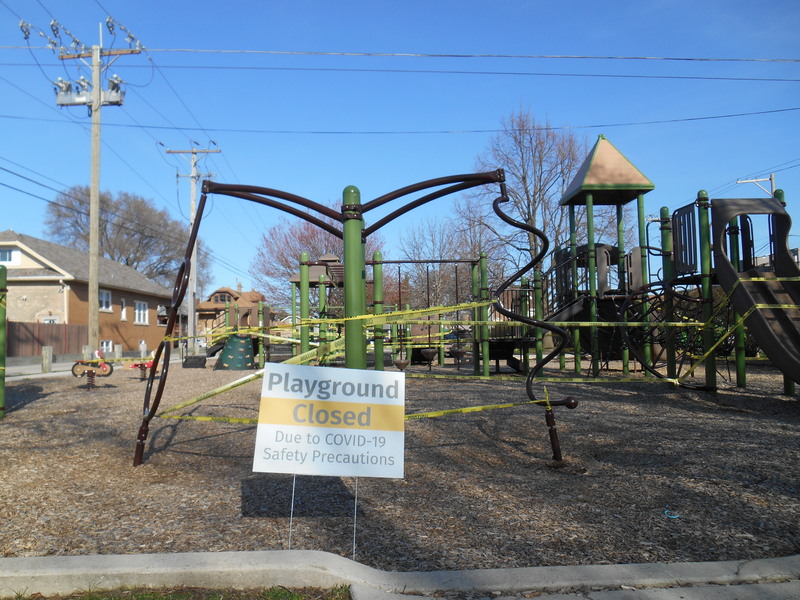 A playground that is blocked off with yellow caution tape. In front of the playground is a sign that says "Playground is Closed". 