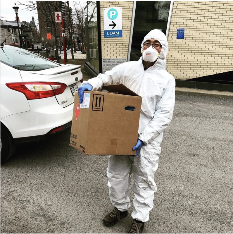 Man in PPE standing outside next to a white car, holding a large cardboard box.