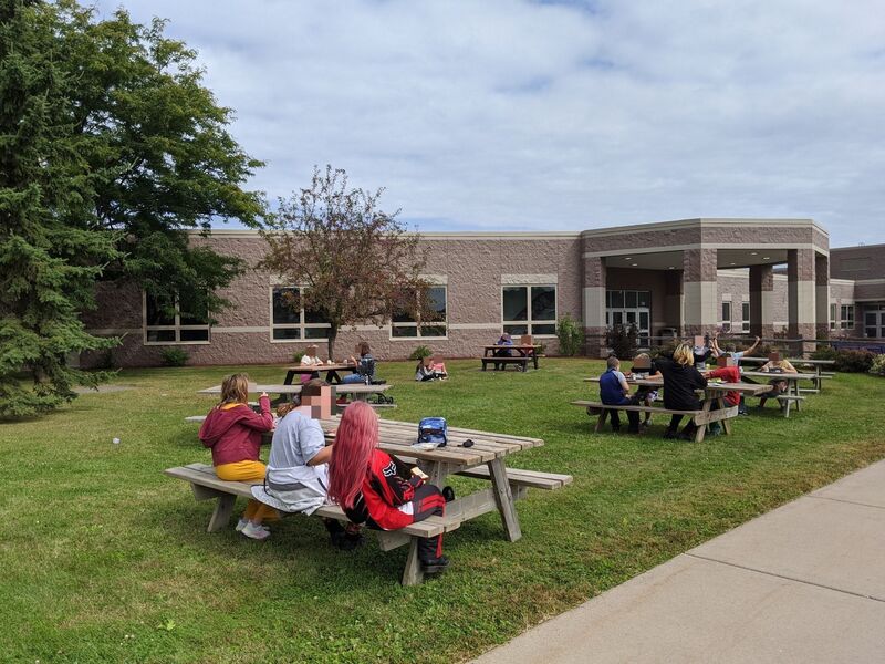 A picture of several groups of people eating lunch at picnic tables outside. 