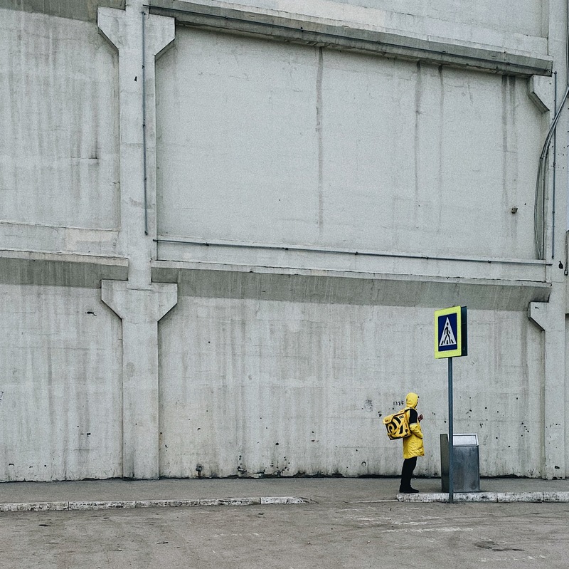 A man in a yellow jacket with a yellow backpack standing by a trash can. 
