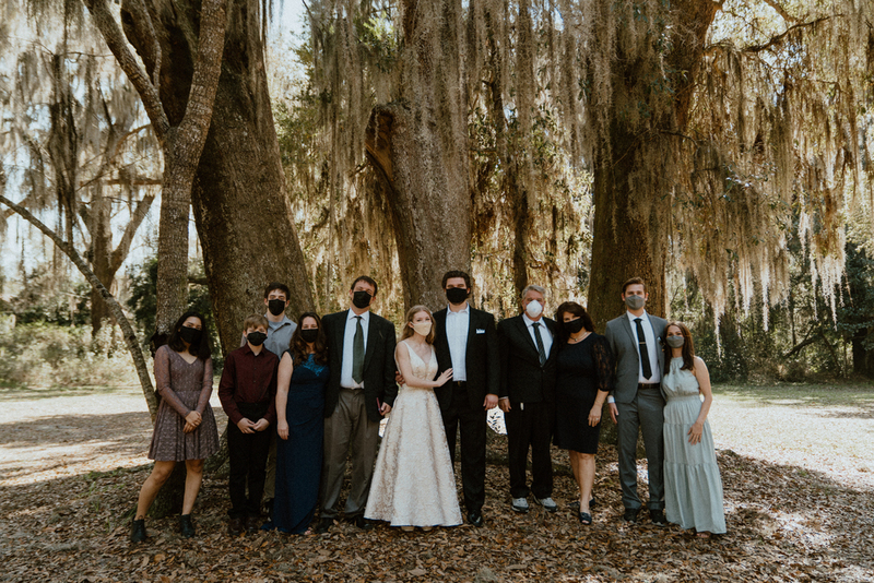 This is a picture of a group of people dressed in formal attire standing outside. All are wearing face masks. This appears to be a wedding photograph. 