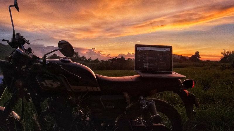 A motorcycle with a laptop on top photographed in front of an orange and pink sunset in a field. 