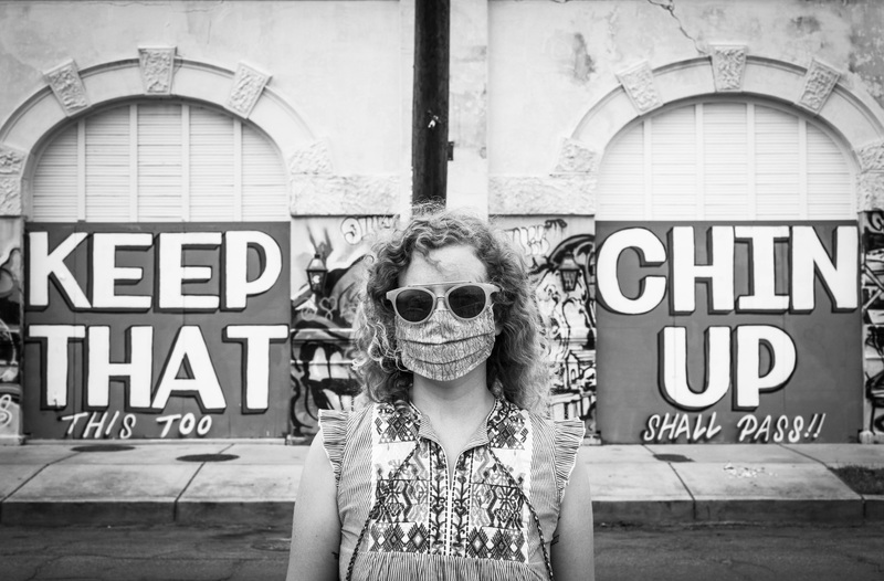 Black and white photo of a woman wearing sunglasses and a face mask in front of a mural that says "Keep that chin up, this too shall pass!"