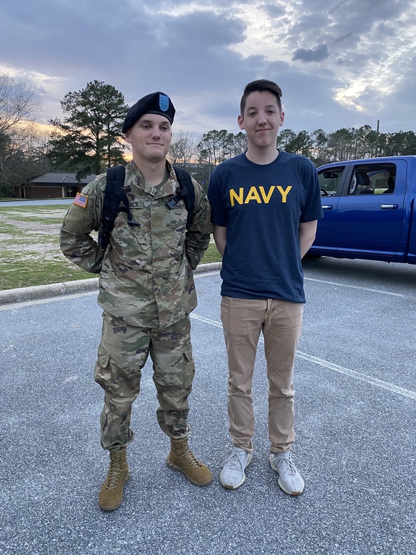 Two people are standing next to each other in a parking lot. The person on the right is in an army uniform wearing a blue beret with a blue logo on it. The person on the right is wearing a navy shirt with the word navy in yellow with khakis. They are both smirking with their hands behind their backs. 