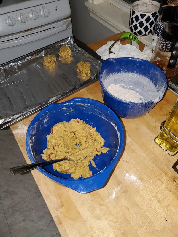 Two blue mixing bowls with baking ingredients. In background is a baking sheet lined with foil with dollops of dough.