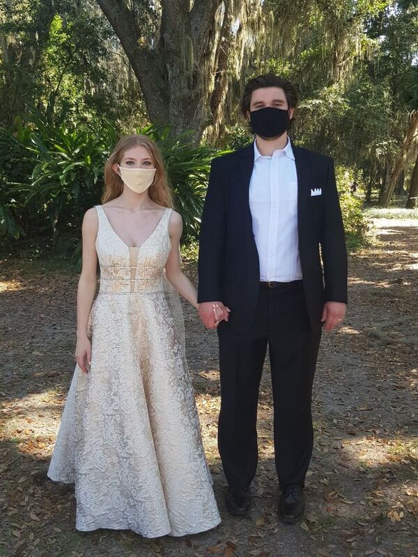 This is a picture of two people dressed in formal attire standing outside. Both are wearing face masks. They appear to be a bride and groom. 