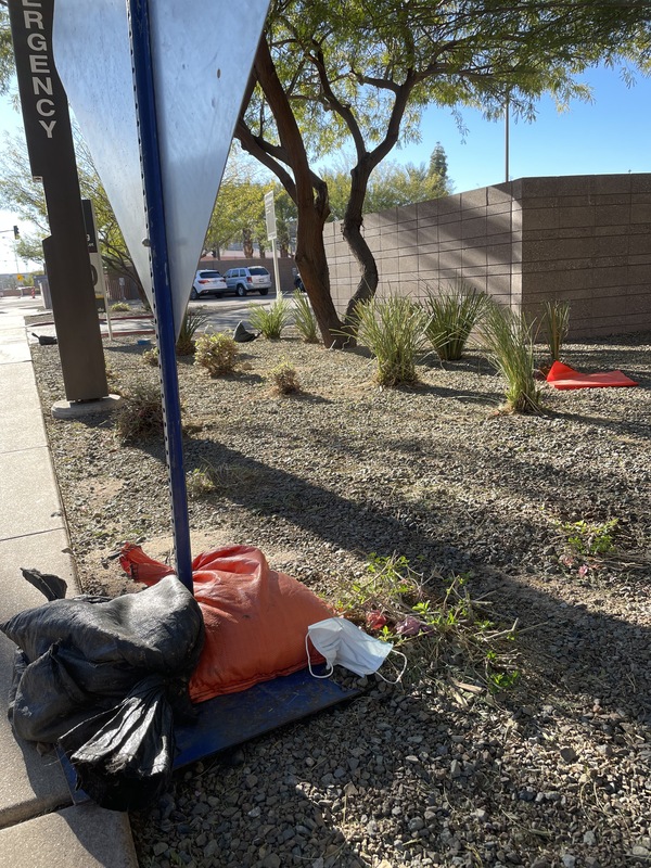 This is a picture of a traffic sign weighed to the ground by an orange sand bag, with a discarded face mask resting at its base. 