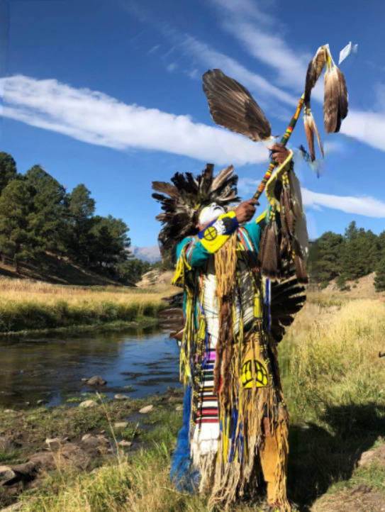 This is a picture of a Native American dressed in traditional ceremonial clothing outdoors by a stream, likely at a powwow. 