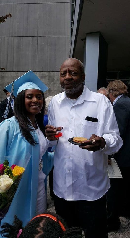 Two people are posing with each other. On the left, the person is wearing a graduation gown and cap and on the right is a person holding a drink in one hand and a plate with cookies in the other. 
