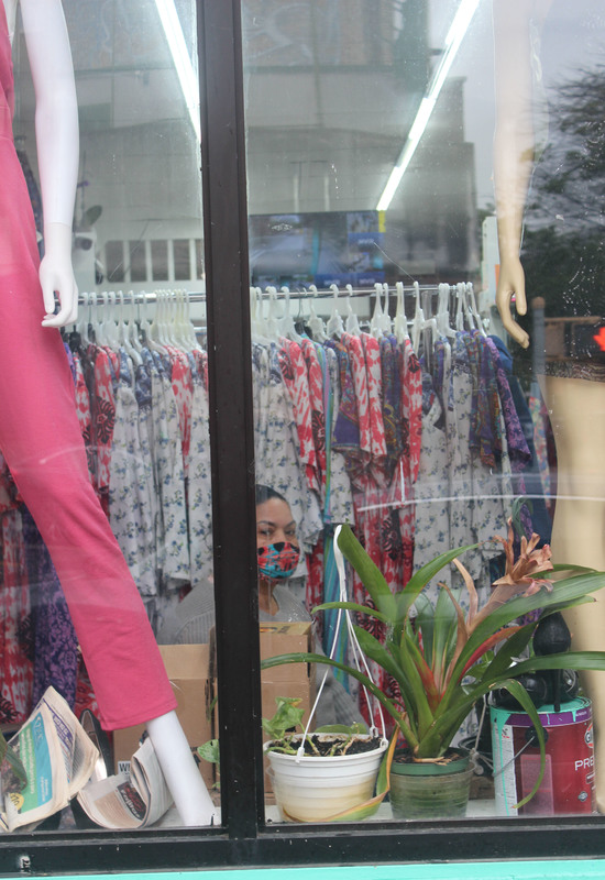 A person with a mask behind windows and plants in front of a rack of clothes.
