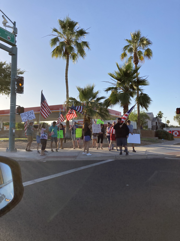 This is a picture of a group of people protesting on the side of a road. They are waving American flags, and are holding signs that protest President Joe Biden. 