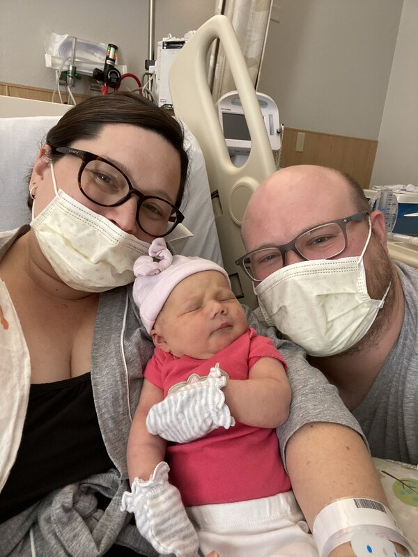 This is a picture of a man and women smiling, both wearing glasses and face masks. The woman is sitting in a hospital bed holding what appears to be a newborn baby.