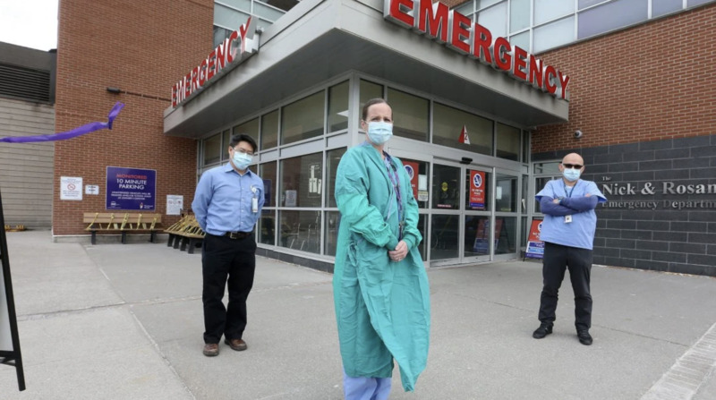 Three hospital workers stand in front of Emergency Room doors.