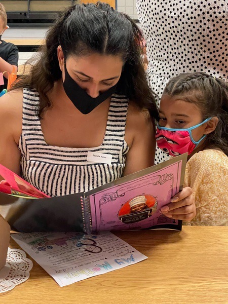 This is a picture of a woman reading from a pink notebook to a younger girl. Both are wearing face masks. 