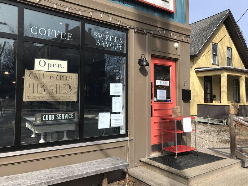 The front of a store has signs taped to the inside of the glass that says: Open. Call to order. Curb Service. In front of the orange door on the right is a red cart. 