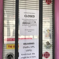 The front of a pink and black glass door that is covered in stickers has two paper signs taped to the inside of a glass that says: Euclid Records in New Orleans is CLOSED. DELIVERIES (FedEx, UPS, USPS): Knock loud. We might be in here. 