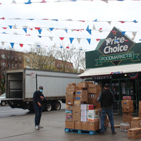 Two workers with masks unload boxes in front of a grocery store.