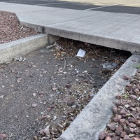 This is a picture of a face mask that has been discarded in the dirt below a drainage area. A concrete walkway extends above it. 