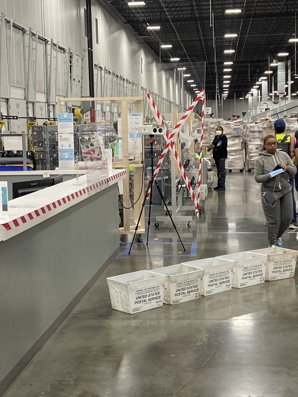 Large Amazon warehouse with the view near a reception like desk with a device on a tripod set up in front of it and Amazon employees lined up in the background.