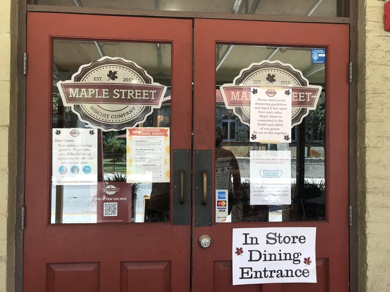 This is a picture taken of the doors leading into the Maple Street Biscuit Company. Several flyers asking people to wear masks and remain socially distant are present on the door.