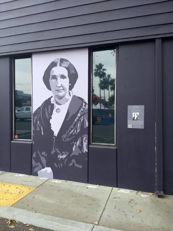 This is a picture taken of a mural that is painted on the side of a building. A woman in antiquated clothing is depicted in black and white.