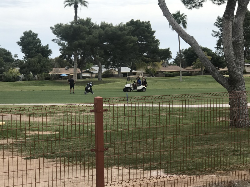 A golf course that has someone golfing while two people look on while sitting in a white golf cart.