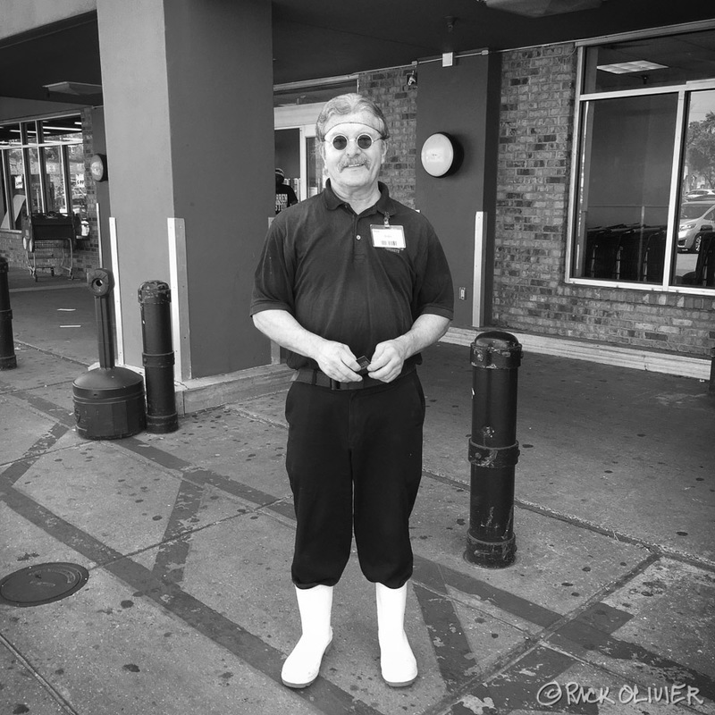 A man is wearing white rain boots with pants tucked into the boots. He is wearing a polo shirt with a name tag attached to it. The man is standing in front of a grocery store. The man is wearing circular sunglasses.