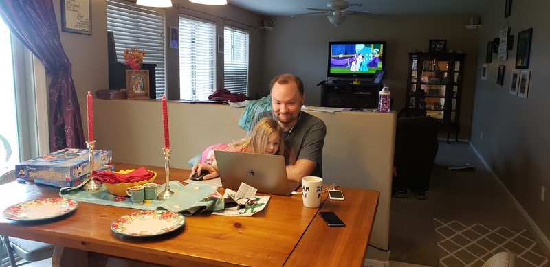 A child and a their Dad are sitting at a wooden dinner table. Dad is sitting in front of a laptop, while his child is leaning over his arm into his lap trying to help. Dad is smirking.