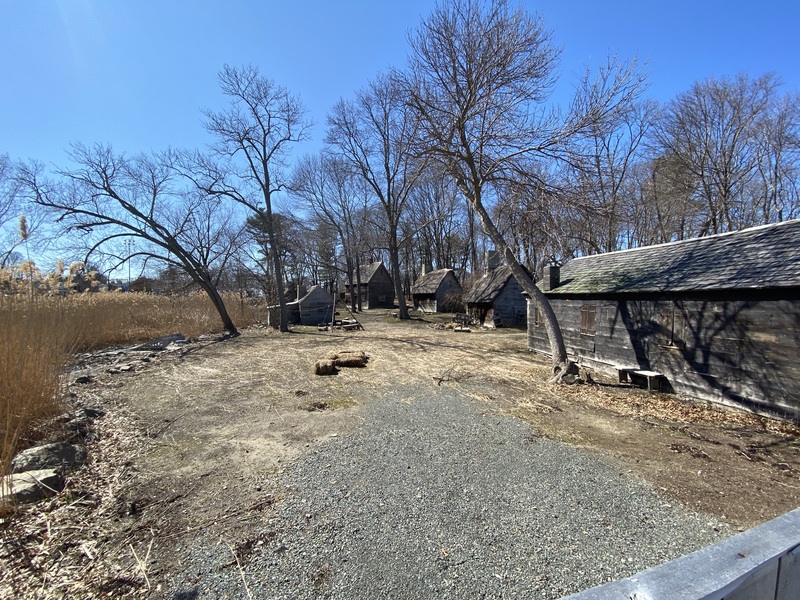 Several old worn buildings surrounded by bare trees, dry grass, and gravel at the Salem Pioneer Village in Salem, Massachusetts.