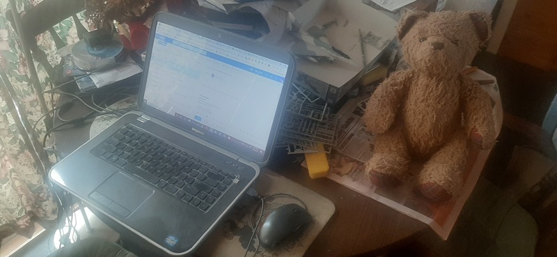 A wooden table with a laptop on top with a teddy bear to the right of it.
