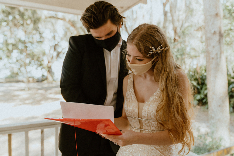 This is a picture of two people dressed in formal attire standing outside. Both are wearing face masks. The woman in the photo seems to be wearing a wedding dress.