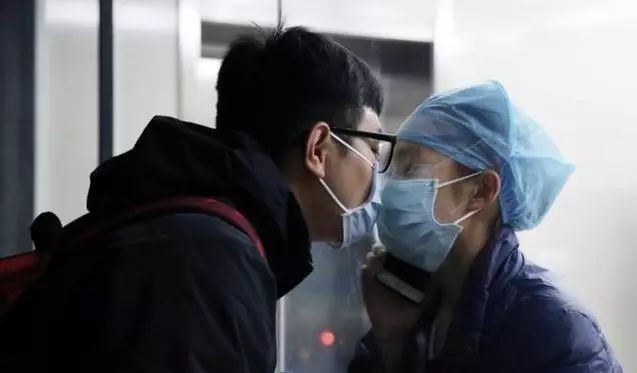 Two people that are wearing face masks and that are separated by a window are kissing each other through the glass. 