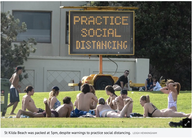 A field of grass has multiple groups of people having picnics and sun bathing. There's an electric sign behind the multiple groups of people that says: PRACTICE SOCIAL DISTANCING.