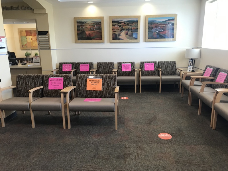 A medical center waiting room featuring chairs blocked off in order to promote social distancing. The signs on the chairs read Do Not Sit Here, please help us practice social distancing and Please Follow The Orange Circles which are on the floor indicating six feet apart for social distancing purposes.