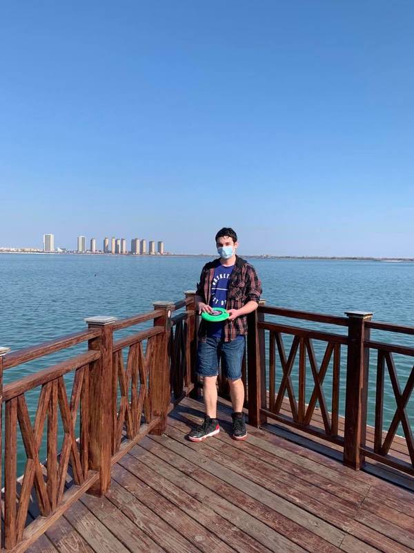 A person is standing on a wooden dock with railings. The person is wearing a plaid jacket with a purple shirt with jean shorts on. The person is holding a green frisbee. Behind the person beyond the water is a row of buildings.