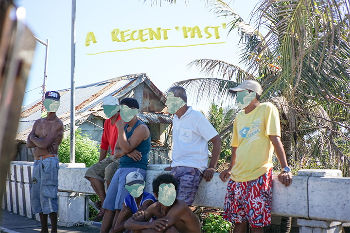 A photograph of seven men leaning on a barrier on the side of a road in front of a shack and palm tree. The faces of the men are blocked out. 