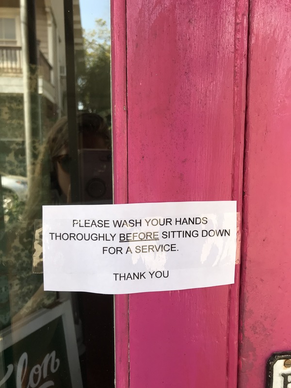 A lamented rectangle sign taped on a bright pink door that says: PLEASE WASH YOUR HANDS THOROUGHLY BEFORE SITTING DOWN FOR A SERVICE. THANK YOU.
