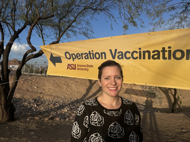 This is a picture taken of a smiling women posing in front of a yellow Arizona State University banner which reads "Operation Vaccination" on it.